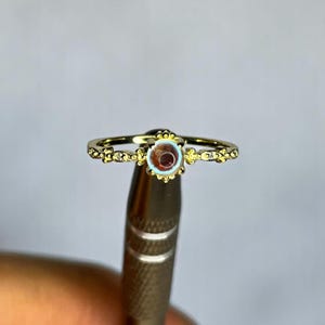 Peut inclure: Une bague dorée avec une pierre précieuse centrale, ronde, brun rougeâtre. La monture de la bague présente des motifs floraux et de points délicats. Le bijou est entouré d'un halo bleu clair. La bague est tenue par un outil en métal.