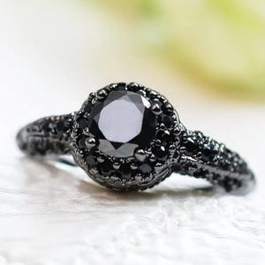 Peut inclure: Une bague en pierre noire avec une pierre centrale ronde et facettée. La bague présente un halo de petites pierres noires autour de la pierre centrale et le long de la bande. La bague est posée sur une surface blanche.