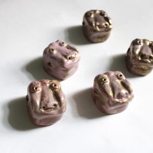 May include: Six ceramic beads in a light purple hue with gold accents. Each bead is cube-shaped and features a stylized face with two eye holes and a nose. The beads are arranged on a white surface.