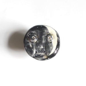 May include: A ceramic face sculpture with a black and white color scheme. The sculpture has a round shape with a face design, featuring two eye sockets, a nose, and a mouth. The face is painted with a black base and white accents.