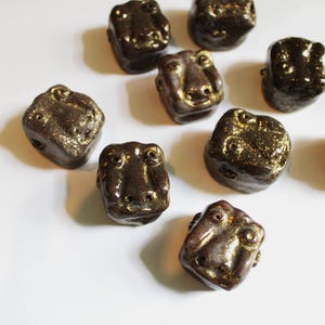 Könnte beinhalten: Eine Sammlung dunkelbrauner, keramikförmiger Froschperlen. Jede Perle hat ein detailliertes Froschgesicht mit sichtbaren Augen und Nasenlöchern. Die Perlen haben eine metallisch goldene Oberfläche und sind auf einer weißen Oberfläche angeordnet.