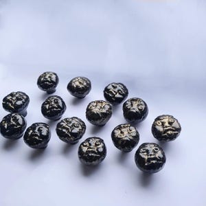 Peut inclure: Ensemble de boutons de meubles sphériques noirs avec un motif de visage en relief détaillé. Les boutons ont un éclat métallique et sont disposés sur une surface blanche. Les visages ont des accents dorés, ajoutant du contraste à la finition sombre.