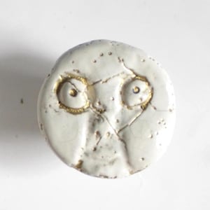 May include: A circular, off-white ceramic button with a face design. It has two eye-shaped openings with gold detailing, and a cracked surface. Small gold dots are scattered across the surface, adding texture and detail.