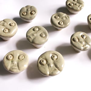 May include: A selection of ceramic cabinet knobs, each with a distinct face design. The knobs are a pale beige hue and display diverse facial expressions, with recessed eyes and mouths. These decorative knobs are perfect for adding a quirky touch to furniture.