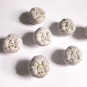 May include: Seven off-white ceramic face-shaped knobs with gold accents. The knobs have a detailed, slightly distressed appearance, with visible facial features. They are arranged on a white surface, casting soft shadows.
