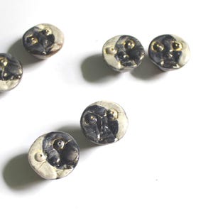 May include: Six handcrafted ceramic buttons with a unique face design. Each button features a cream-coloured base with a dark grey face and gold accents. These decorative buttons are ideal for sewing and craft projects.