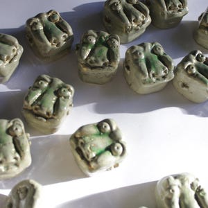 Könnte beinhalten: Eine Sammlung kleiner, handgefertigter Keramikskulpturen. Jede Skulptur ist ein kubusförmiger Kopf mit einem Gesicht, bemalt in Grün- und Grautönen. Die Skulpturen sind auf einer weißen Oberfläche angeordnet.