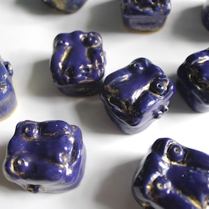 May include: A collection of ceramic frog figurines. Each frog is a deep blue with gold accents, featuring a glossy finish. The frogs have detailed facial features and a whimsical design, perfect for home decor or as unique collectibles.