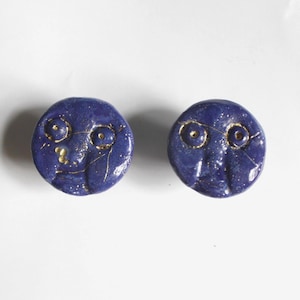 May include: Two round, dark blue ceramic objects with gold accents. Each has a face design with gold-lined eyes and a mouth. The objects are on a white background. The ceramic pieces have a textured surface.