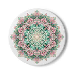 May include: A round, white coaster featuring a detailed mandala design. The mandala is rendered in shades of pink, green, and gold, with intricate floral and geometric patterns radiating from the center. The coaster has a smooth, matte finish.