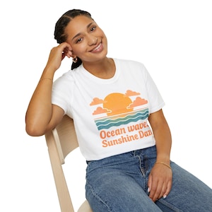 Puede incluir: Camiseta blanca con un diseño de puesta de sol: sol naranja, nubes melocotón y olas oceánicas azules y verdes. El texto "Ocean wave Sunshine Daze" está impreso en naranja. La camiseta se combina con unos vaqueros azules.