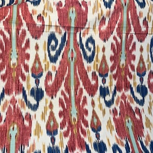 May include: A fabric featuring a repeating ikat pattern in shades of deep red, navy blue, and light teal on an off-white background. The design consists of elongated, symmetrical floral-like motifs with blurred edges and vertical orientation.