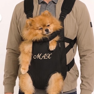 May include: A tan Pomeranian dog is nestled in a black pet carrier with the name "MAX" embroidered on the front. The carrier has adjustable straps and is worn by a person wearing a tan jacket.