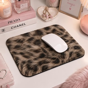 Könnte beinhalten: Ein rechteckiges Mauspad mit einem braun-schwarzen Leopardenmuster und einer glatten, flachen Oberfläche. Eine weiße kabellose Computermaus befindet sich in der Mitte des Pads, das abgerundete Ecken und einen dünnen schwarzen Rand aufweist.