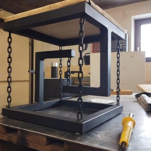 Puede incluir: Una mesa única, hecha a mano, con estructura de metal negro y tapa de madera. La mesa parece flotar, suspendida por cadenas. El diseño incorpora formas geométricas y elementos industriales, creando una estética moderna.