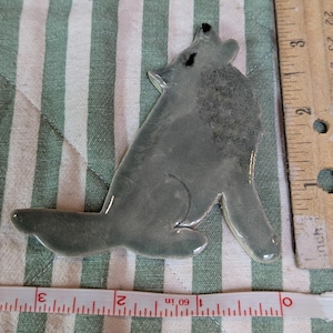 May include: A ceramic howling wolf figurine in shades of gray, with black accents. The wolf is in a sitting position, with its head tilted back. The figurine is approximately 3 inches long.