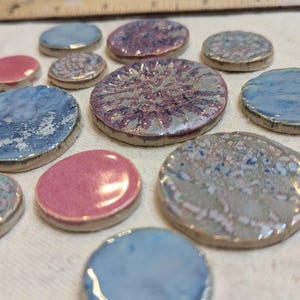 Puede incluir: Colección de baldosas de cerámica ovaladas en varios colores, incluyendo rosa, azul y morado. Las baldosas tienen un acabado brillante y un aspecto texturizado, con algunos diseños moteados. Los bordes son de un color más oscuro.