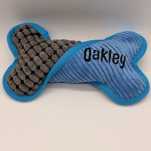 Puede incluir: Un juguete de peluche con forma de hueso para perros, azul y gris. La sección central es gris con un patrón texturizado, mientras que los extremos son de pana azul con el nombre "Oakley" bordado en negro. El juguete tiene un ribete azul.
