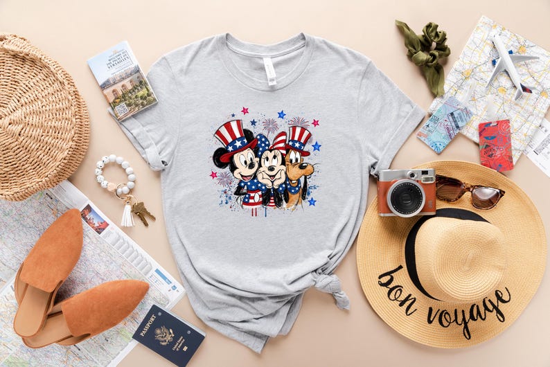 K&ouml;nnte beinhalten: Ein hellgraues Kurzarm-T-Shirt mit einer Grafik von Mickey Maus, Minnie Maus und Pluto, die rot-wei&szlig;-blaue patriotische Zylinder und sternenbedruckte Kleidung tragen, umgeben von Illustrationen blauer und roter Feuerwerke.