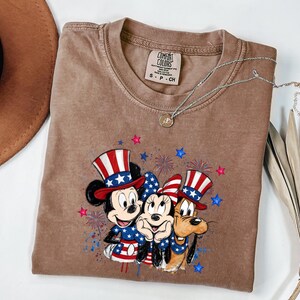 K&ouml;nnte beinhalten: Ein braunes Kurzarm-T-Shirt mit einer Grafik von Mickey Maus, Minnie Maus und Pluto, die patriotische rot-wei&szlig;-blaue Zylinder und Outfits tragen, umgeben von Feuerwerks- und Sternillustrationen. Das T-Shirt hat ein Comfort Colors Etikett.