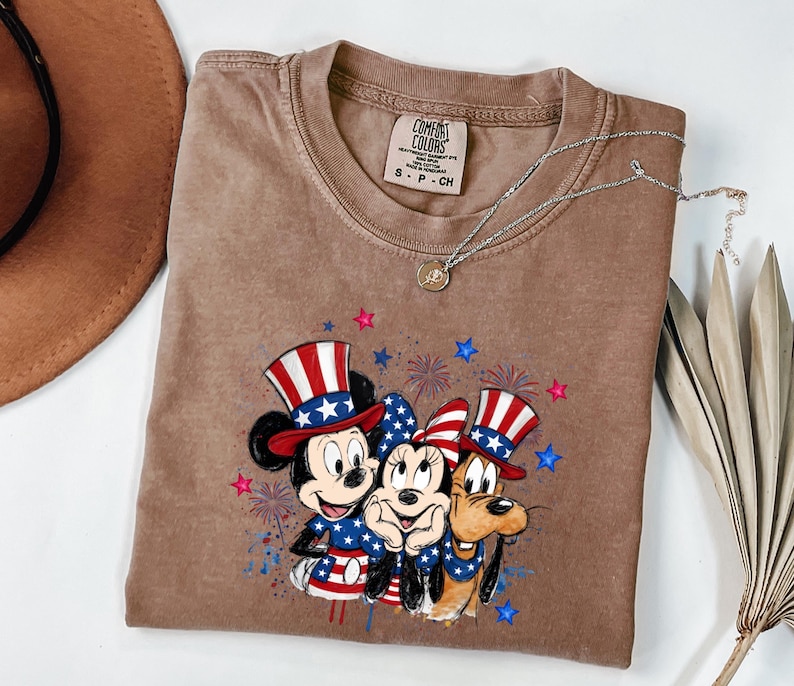 K&ouml;nnte beinhalten: Ein braunes Kurzarm-T-Shirt mit einer Grafik von Mickey Maus, Minnie Maus und Pluto, die patriotische rot-wei&szlig;-blaue Zylinder und Outfits tragen, umgeben von Feuerwerks- und Sternillustrationen. Das T-Shirt hat ein Comfort Colors Etikett.
