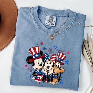 K&ouml;nnte beinhalten: Ein hellblaues T-Shirt mit Rundhalsausschnitt und einem Grafikdruck von drei Zeichentrickfiguren, die patriotische Zylinder und Outfits in Rot, Wei&szlig; und Blau tragen, umgeben von roten und blauen Sternen sowie Feuerwerksmotiven.