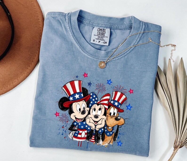 K&ouml;nnte beinhalten: Ein hellblaues T-Shirt mit Rundhalsausschnitt und einem Grafikdruck von drei Zeichentrickfiguren, die patriotische Zylinder und Outfits in Rot, Wei&szlig; und Blau tragen, umgeben von roten und blauen Sternen sowie Feuerwerksmotiven.