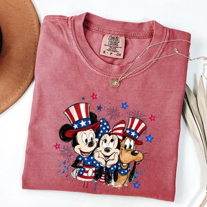 K&ouml;nnte beinhalten: Ein altrosa T-Shirt mit kurzen &Auml;rmeln und einer Grafik von Mickey Mouse, Minnie Mouse und Pluto, die patriotische rot-wei&szlig;-blaue H&uuml;te und Outfits tragen, umgeben von blauen und rosa Sternen und Feuerwerksmotiven, mit einem Comfort Colors Etikett.