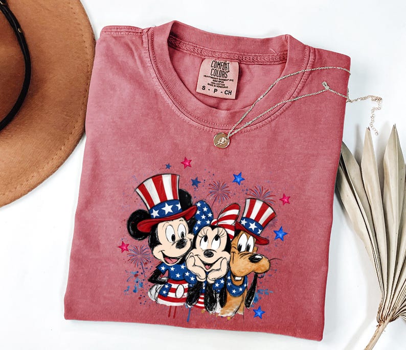 K&ouml;nnte beinhalten: Ein altrosa T-Shirt mit kurzen &Auml;rmeln und einer Grafik von Mickey Mouse, Minnie Mouse und Pluto, die patriotische rot-wei&szlig;-blaue H&uuml;te und Outfits tragen, umgeben von blauen und rosa Sternen und Feuerwerksmotiven, mit einem Comfort Colors Etikett.