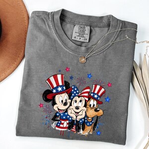K&ouml;nnte beinhalten: Ein anthrazitfarbenes Kurzarm-T-Shirt mit einer Grafik von Mickey Maus, Minnie Maus und Pluto, die patriotische rot-wei&szlig;-blaue Zylinder und Outfits tragen, akzentuiert durch Feuerwerk und Sterne. Das Innenetikett zeigt den Schriftzug Comfort Colors.