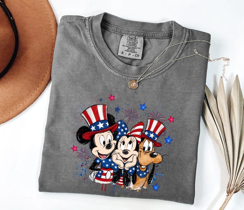 K&ouml;nnte beinhalten: Ein anthrazitfarbenes Kurzarm-T-Shirt mit einer Grafik von Mickey Maus, Minnie Maus und Pluto, die patriotische rot-wei&szlig;-blaue Zylinder und Outfits tragen, akzentuiert durch Feuerwerk und Sterne. Das Innenetikett zeigt den Schriftzug Comfort Colors.