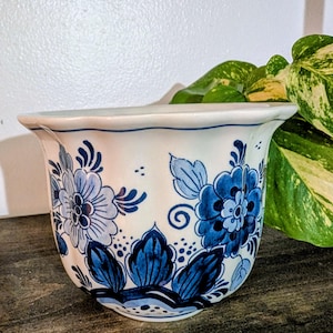 May include: A white ceramic planter with a blue floral design. The planter has a scalloped rim and is decorated with stylized blue flowers and leaves. The design is reminiscent of Delftware pottery. The planter is suitable for indoor or outdoor use.