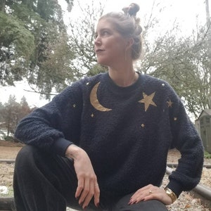 Könnte beinhalten: Marineblauer Pullover mit einem goldenen Halbmond- und Sterndesign. Der Pullover hat ein strukturiertes Aussehen und verfügt über kleine goldene Sternakzente. Die Ärmel und der Halsausschnitt haben einen dunkelblauen Rippenbesatz. Der Pullover ist ein gemütliches und stilvolles Kleidungsstück.