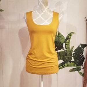 Pode incluir: Top sem mangas amarelo mostarda com decote redondo, exposto em um manequim branco. A parte superior sem mangas tem uma silhueta ajustada e é feita de um material macio e elástico. O fundo apresenta uma parede branca e um desenho de folhas tropicais pintadas.