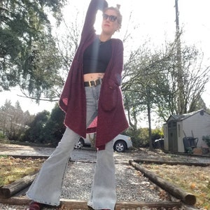 May include: A woman wearing a maroon cardigan with a black crop top and light blue flared jeans. She is wearing sunglasses and a gold belt.