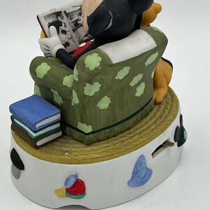 Może przedstawiać: Ceramiczna figurka przedstawiająca Myszka Miki czytającego książkę, siedzącego w zielonym fotelu z białymi kropkami. Kubuś Puchatek siedzi obok Myszki Miki. Stos książek znajduje się na podstawie. Figurka stoi na białej podstawie z kolorowymi wzorami.