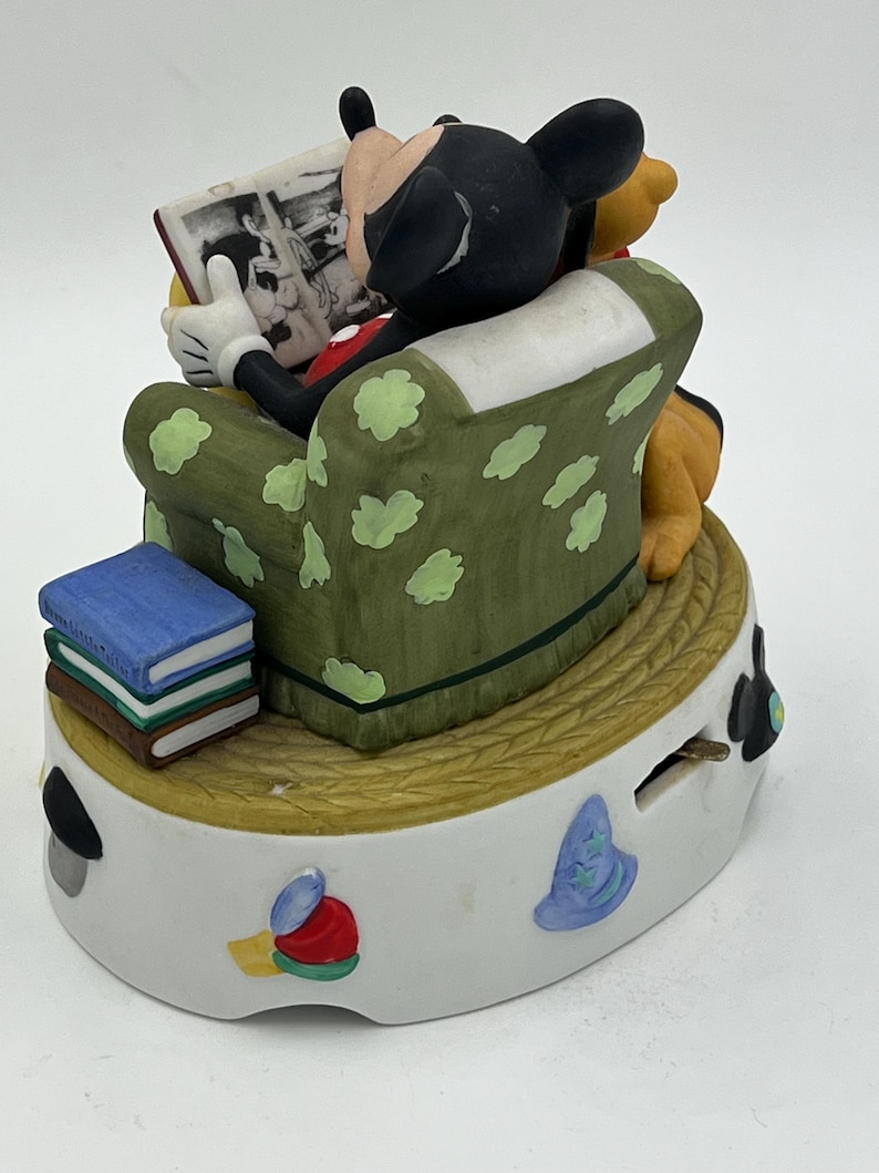 Może przedstawiać: Ceramiczna figurka przedstawiająca Myszka Miki czytającego książkę, siedzącego w zielonym fotelu z białymi kropkami. Kubuś Puchatek siedzi obok Myszki Miki. Stos książek znajduje się na podstawie. Figurka stoi na białej podstawie z kolorowymi wzorami.