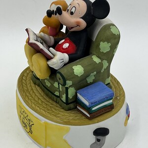 Może przedstawiać: Ceramiczna figurka Myszki Miki i Pluto czytających książkę. Miki siedzi w zielonym fotelu w białe grochy, trzymając książkę. Pluto jest obok niego. Na podstawie widnieją napisy "Mickey Mouse" i "Walt Disney World".