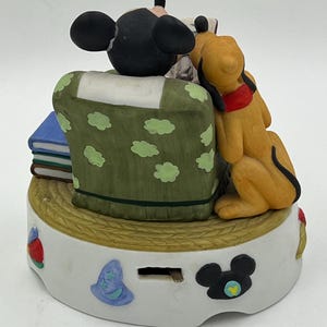 Może przedstawiać: Ceramiczna figurka przedstawiająca Myszka Miki i Pluto. Miki siedzi na zielonym krześle w białe kropki, a Pluto opiera się o krzesło. Podstawa jest biała z elementami dekoracyjnymi, w tym sylwetką Myszki Miki.