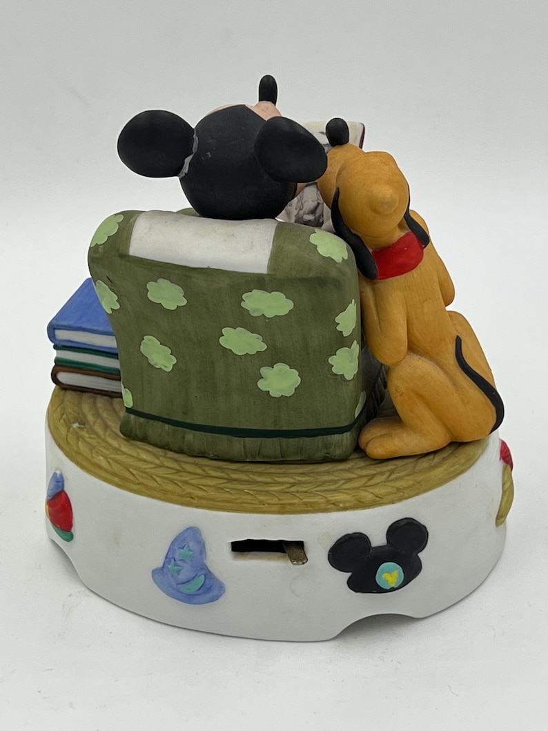 Może przedstawiać: Ceramiczna figurka przedstawiająca Myszka Miki i Pluto. Miki siedzi na zielonym krześle w białe kropki, a Pluto opiera się o krzesło. Podstawa jest biała z elementami dekoracyjnymi, w tym sylwetką Myszki Miki.
