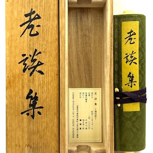 May include: An open wooden box with a scroll and a label inside. The box has Japanese characters on the front. The scroll is green with yellow accents and purple ties. The label has Japanese text.