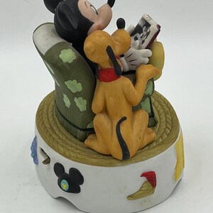 Może przedstawiać: Ceramiczna figurka przedstawiająca Myszka Miki, Pluto i zdjęcie. Myszka Miki jest czarno-biały, Pluto jest ż&oacute;łty z czerwoną obrożą, a podstawa jest biała z elementami dekoracyjnymi. Figurka ma okrągłą podstawę o fakturze liny.