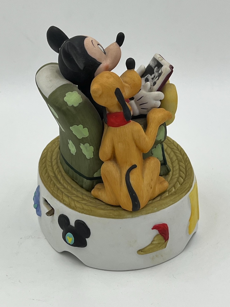 Może przedstawiać: Ceramiczna figurka przedstawiająca Myszka Miki, Pluto i zdjęcie. Myszka Miki jest czarno-biały, Pluto jest ż&oacute;łty z czerwoną obrożą, a podstawa jest biała z elementami dekoracyjnymi. Figurka ma okrągłą podstawę o fakturze liny.