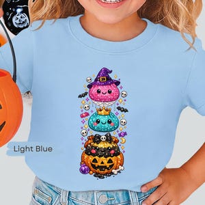 Peut inclure: Un t-shirt bleu clair avec un motif vertical représentant trois personnages empilés et scintillants : un rose avec un chapeau de sorcière violet, un bleu sarcelle avec une couronne dorée et un noir dans une citrouille orange avec un nœud tête de mort.