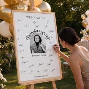 Op de afbeelding: Een wit rechthoekig welkomstbord voor een verjaardagsfeest op een houten ezel met de tekst Welcome to Hannah's 30th Birthday Party September 26th, 2026, een centraal zwart-witportret van een persoon en diverse handgeschreven handtekeningen.