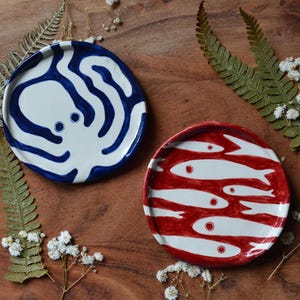 Op de afbeelding: Twee keramische borden met mariene ontwerpen. Eén bord is wit met een blauw octopusontwerp. Het andere bord is rood met witte vissen. De borden liggen op een houten oppervlak met varens en witte bloemen.