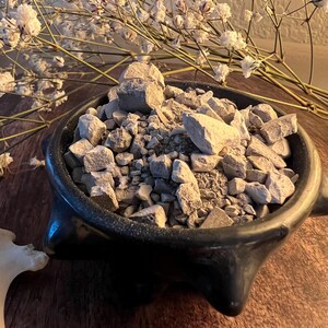 May include: A black ceramic bowl filled with light gray, irregularly shaped stones. The bowl has three decorative feet and is set on a wooden surface. White dried flowers are in the background.
