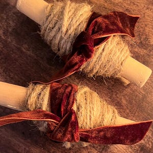 May include: Two wooden spools wrapped with natural twine and adorned with a deep red velvet ribbon. The spools are arranged on a dark wooden surface, creating a rustic and elegant aesthetic. Ideal for craft projects or decorative use.