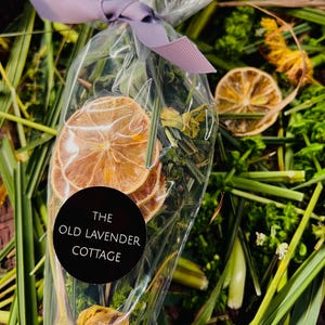 May include: A clear cellophane bag tied with a lavender ribbon, filled with dried citrus slices and herbs. A black circular label with white text reads "THE OLD LAVENDER COTTAGE". The background features green foliage and dried flowers.
