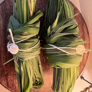 May include: Two bundles of green lemongrass, wrapped and tied with white string and a button closure. The bundles are arranged on a round, wooden surface. The fresh green leaves are the focal point of the image.